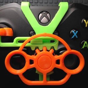 Op de afbeelding: Close-up van een zwarte Xbox-controller met een neon groen en oranje stuurwielbevestiging. De controller heeft gekleurde knoppen en een centraal Xbox-logo. De bevestiging lijkt 3D-geprint te zijn.