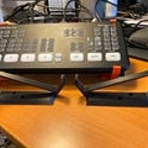 May include: Two black metal brackets with a rectangular base and a vertical support, designed to hold equipment. The brackets are positioned on a wooden surface, with a black control panel in the background.