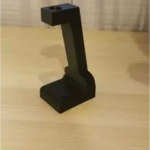 May include: A black stand with a rectangular base and a cylindrical hole at the top. The stand is made of a solid material and is positioned on a light-coloured wooden surface.