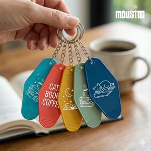 May include: A set of five colorful keychains with silver chain links. Each keychain has a different color and design: teal with books, red with "CAT BOOK COFFEE", yellow with a moon and book, green with coffee and books, and blue with a cat on a book.