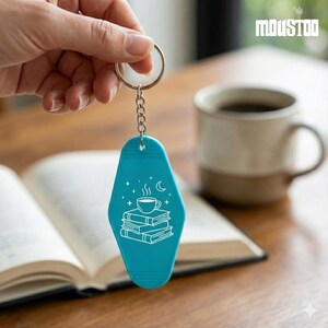 May include: Teal keychain with a silver ring and chain. The keychain features a white graphic of a coffee cup on a stack of books, with stars and a crescent moon. The word "MOUSTOO" is in the upper right corner.