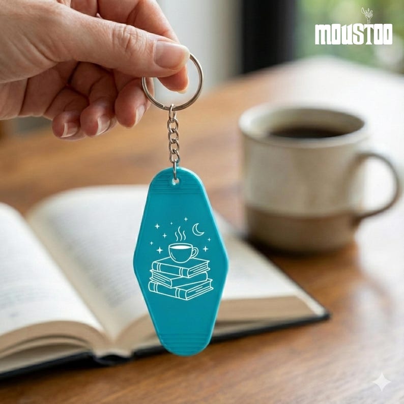 May include: Teal keychain with a silver ring and chain. The keychain features a white graphic of a coffee cup on a stack of books, with stars and a crescent moon. The word "MOUSTOO" is in the upper right corner.