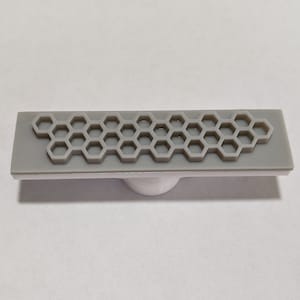 May include: A gray rectangular rubber stamp with a honeycomb pattern. The stamp is mounted on a white plastic base.