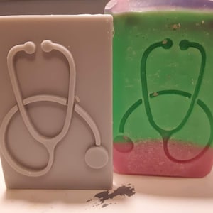 May include: Two soap bars featuring a stethoscope design. One bar is grey with a white stethoscope design. The other bar is green and pink with a white stethoscope design.