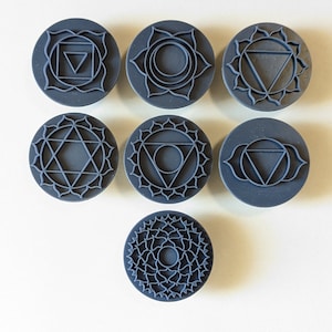 Puede incluir: Siete sellos circulares de color gris oscuro con intrincados diseños geométricos que representan los chakras. Cada sello presenta un símbolo único dentro de un borde floral o geométrico. Los sellos están dispuestos sobre una superficie blanca.