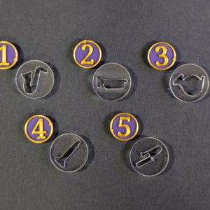 May include: A selection of game tokens and clear acrylic cutouts. The tokens are purple with gold edging and numbered 1 to 5. The cutouts depict musical instruments: saxophone, tuba, trumpet, clarinet, and trombone.