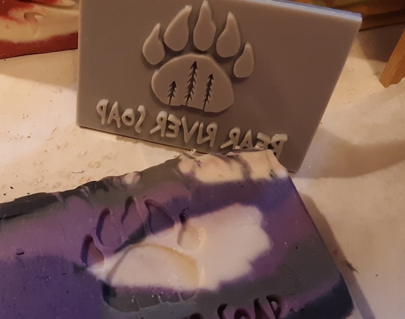 May include: A bar of purple, white, and gray soap with a paw print design. The soap is labeled "Bear River Soap".