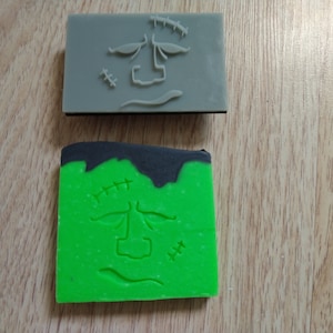 May include: A green and black soap bar with a Frankenstein face design. The soap bar is next to a gray mold with the same design.