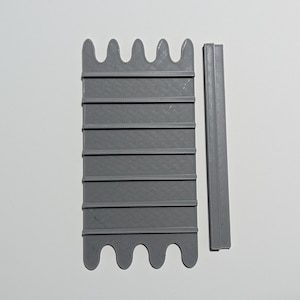 Puede incluir: Un molde de plástico gris impreso en 3D con una serie de crestas y un borde ondulado. También se incluye una tira de plástico gris separada, también impresa en 3D.