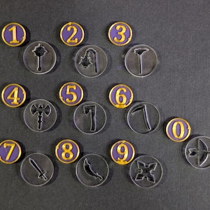 May include: A collection of clear acrylic game tokens with gold-colored rims and purple centers. Each token features a number from 0 to 9 in gold, and a weapon or symbol. The tokens are arranged on a dark gray surface.
