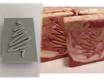 May include: A gray silicone mold with a Christmas tree design. The mold is used to create soap bars with a raised Christmas tree design. The soap bars are red, white, and beige with a swirl pattern.