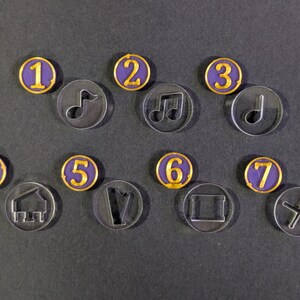 Puede incluir: Una colección de fichas de acrílico transparente con varios símbolos y números. Las fichas numeradas tienen un centro morado con borde dorado. Los símbolos incluyen notas musicales, una casa, una forma de V, un cuadrado y una cruz.
