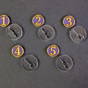 以下が含まれることがあります： ゲームトークンと透明アクリルカットアウトのコレクション。トークンは紫と金色の縁取りで、1から5まで番号が振られています。透明アクリルカットアウトはギターの形をしています。アイテムは濃い灰色の表面に配置されています。