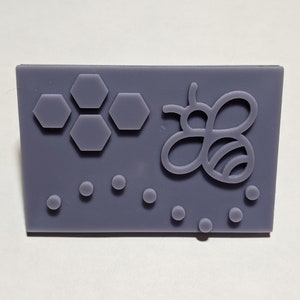 Peut inclure: Un moule en silicone violet avec un motif d'abeille et de nid d'abeille. Le moule est rectangulaire et a une surface texturée.