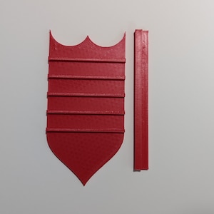 May include: A red, shield-shaped object with a textured surface and a decorative top, alongside a matching rectangular piece. The shield has horizontal lines and a pointed bottom. The objects are likely components of a larger item.