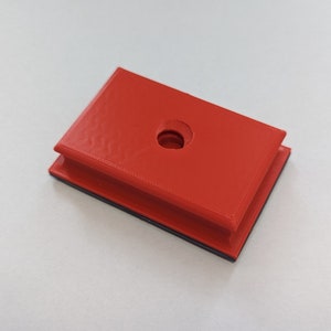 May include: A red plastic block with a round hole in the center. The block has a black border around the edges.
