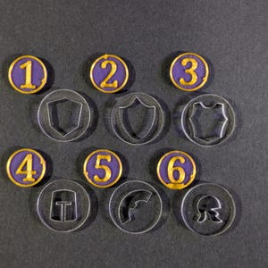 Puede incluir: Una colección de fichas y marcadores de juego. Seis fichas numeradas moradas y doradas del 1 al 6 están dispuestas con seis fichas acrílicas transparentes con diseños de escudo, armadura y casco. Los artículos están sobre una superficie gris oscuro.