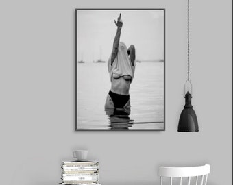 May include: Black and white photograph of a person standing in the water with their shirt pulled over their head, giving the middle finger.
