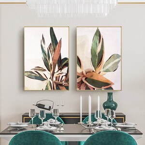 May include: Two framed watercolor paintings of green and brown tropical leaves with white accents. The paintings are hanging on a white wall above a dining table with green chairs.