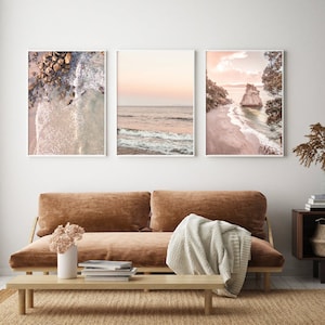 May include: Three framed prints of beach scenes with pink and blue hues. The first print shows a rocky beach with waves crashing on the shore. The second print shows a wide view of the ocean with waves and a pink sky. The third print shows a beach with a rocky outcropping and a pink sky.
