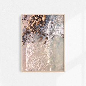 May include: An aerial view of a rocky shoreline with waves crashing on the sandy beach. The water is a light blue and the rocks are a dark brown.