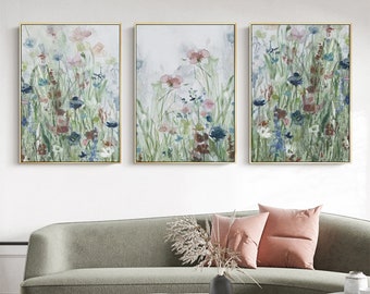May include: Three framed watercolor paintings depicting a field of wildflowers in shades of blue, pink, and green. The paintings are arranged in a row on a white wall.