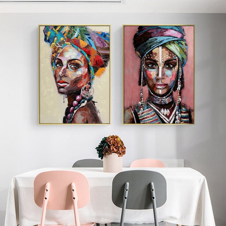 May include: Two framed paintings of women with colorful head wraps and jewelry. The women are depicted with bold, expressive features and intricate details. The paintings are set against a white background.
