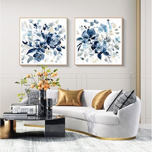 May include: Two square framed watercolor paintings of blue floral bouquets on a white background. The paintings are hanging on a white wall above a white sofa with gold accents.