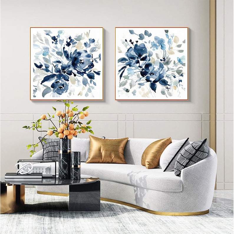 May include: Two square framed watercolor paintings of blue floral bouquets on a white background. The paintings are hanging on a white wall above a white sofa with gold accents.