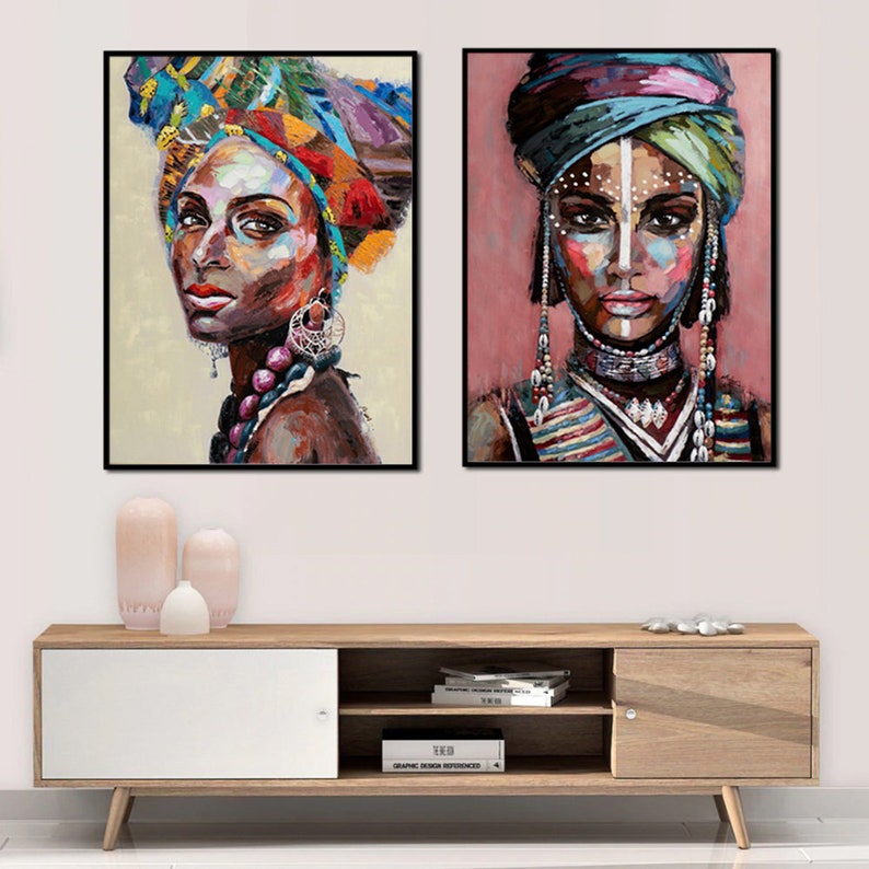 May include: Two colorful paintings of women wearing head wraps and traditional jewelry. The paintings are framed in black and set against a white wall.