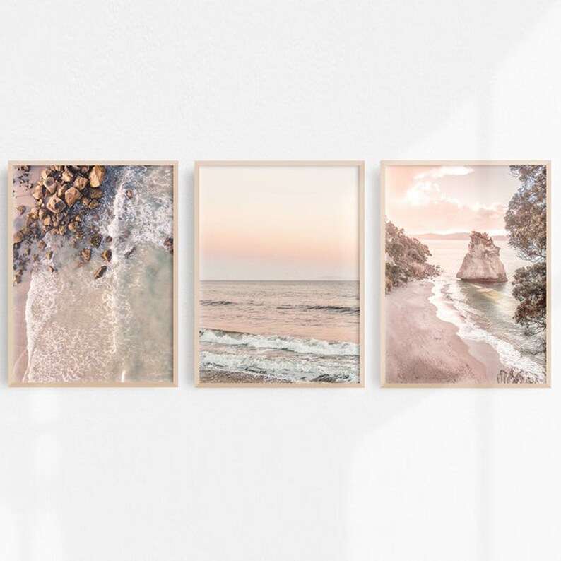 May include: Three framed prints of seascapes with soft, pastel colors. The first print shows a close-up of foamy waves and rocks. The second print shows a wide view of the ocean with a pink sky. The third print shows a rocky beach with a small island in the distance.