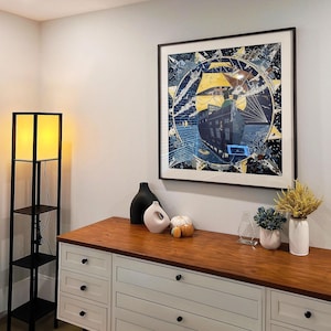 May include: A square framed wall art print featuring a dark blue and yellow nautical scene with a ship, surrounded by a star-shaped border. The artwork is displayed above a wooden dresser with white drawers, next to a tall black floor lamp with a yellow light.