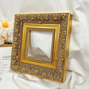May include: A gold picture frame with ornate detailing. The frame has a brown inner border and a gold outer border. The frame is square shaped.