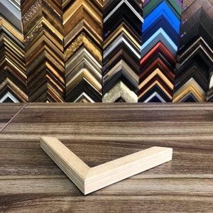May include: A light-coloured wooden picture frame corner rests on a wooden surface. The corner is angled. In the background, a variety of picture frame samples in different colours and styles are displayed, including gold, black, and silver.
