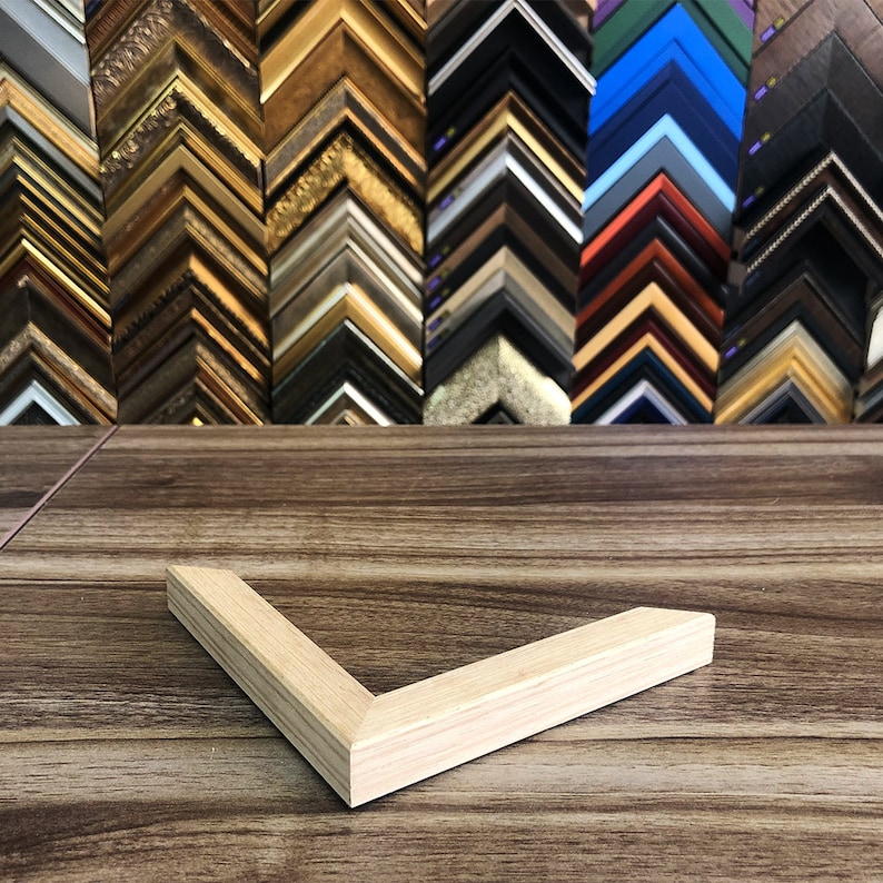 May include: A light-coloured wooden picture frame corner rests on a wooden surface. The corner is angled. In the background, a variety of picture frame samples in different colours and styles are displayed, including gold, black, and silver.