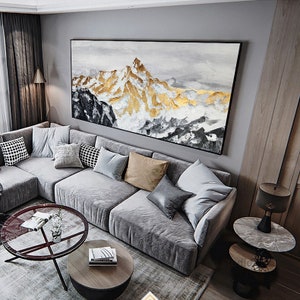 May include: A gray sectional sofa with multiple pillows in a living room setting. A large abstract painting of a mountain range with gold accents hangs on the wall above the sofa. The painting is framed in black.