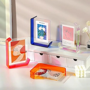 May include: Three acrylic photo frames in different colours, blue, pink, and orange. The frames are on a white shelf with drawers. The frames are holding photos of flowers, a line drawing of a face, and a line drawing of a face with text.