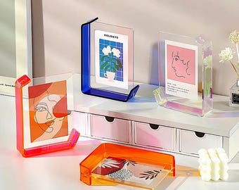May include: Three acrylic photo frames in different colors, blue, pink, and orange. The frames are on a white shelf with drawers. The frames are holding photos of flowers, a line drawing of a face, and a line drawing of a face with text.