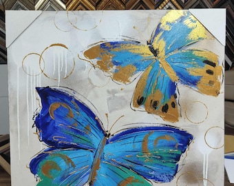 May include: A square canvas art piece featuring two vibrant butterflies. The butterflies are painted with shades of blue, teal, and gold against a white and gray background. The artwork has a modern, abstract style with gold accents and circular patterns.