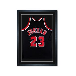 May include: A black basketball jersey with red and white trim, featuring the name "JORDAN" and the number "23" on the back. The jersey is framed in a black frame.