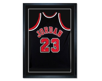 May include: A black basketball jersey with red and white trim, featuring the name "JORDAN" and the number "23" on the back. The jersey is framed in a black frame.