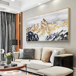 May include: Abstract painting of a mountain range with gold and white accents. The painting is framed in a gold frame and is hanging on a white wall in a living room setting.