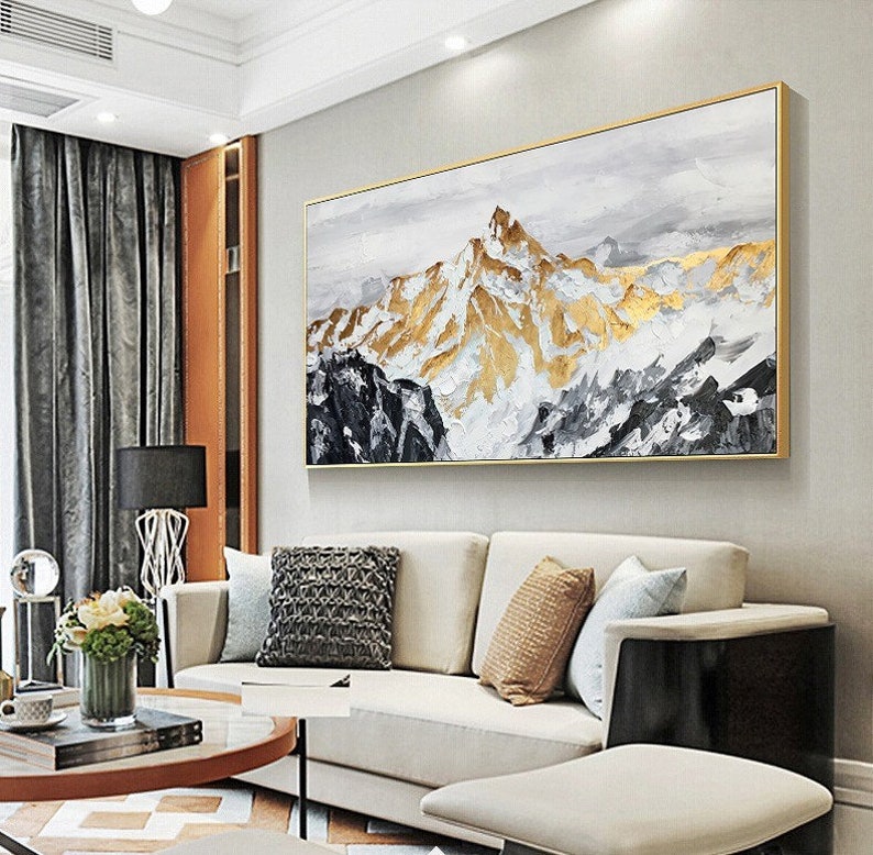 May include: Abstract painting of a mountain range with gold and white accents. The painting is framed in a gold frame and is hanging on a white wall in a living room setting.