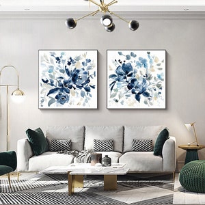 May include: Two square framed watercolor paintings of blue floral bouquets on a white background. The paintings are hanging on a white wall above a white couch with green and black patterned throw pillows.