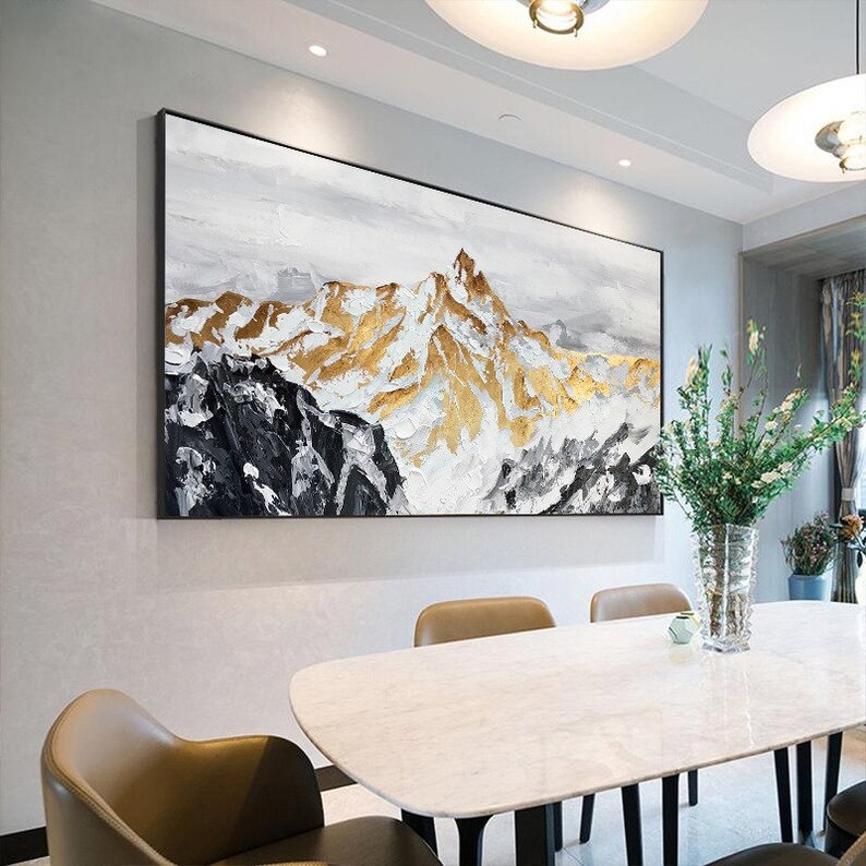 May include: Abstract painting of a mountain range with gold and white accents. The painting is framed in black and hangs on a white wall above a dining table.