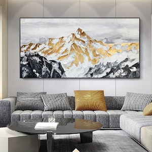 May include: Abstract painting of a mountain range with gold and white accents. The painting is framed and hanging on a wall above a gray sofa.