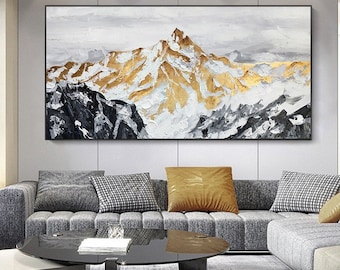 May include: Abstract painting of a mountain range with gold and white accents. The painting is framed and hanging on a wall above a gray sofa.