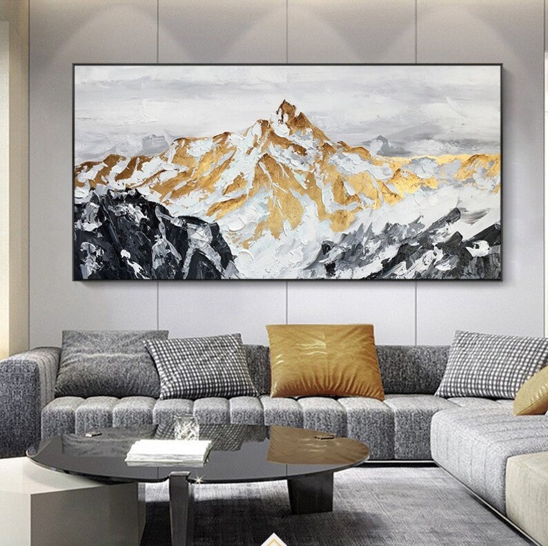 May include: Abstract painting of a mountain range with gold and white accents. The painting is framed and hanging on a wall above a gray sofa.
