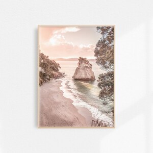 May include: A pink and white photograph of a rocky island in the ocean. The island is surrounded by a sandy beach and clear blue water. The sky is a soft pink and white, with clouds.