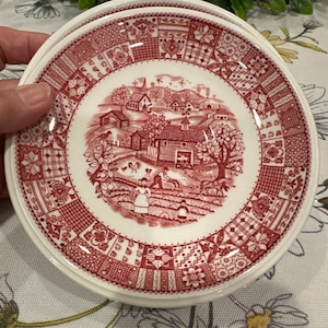 May include: Two white ceramic plates with a red and white toile design. The plates show a rural landscape scene and a patchwork border. The plates are held by a hand. Each plate measures approximately 20 cm in diameter.